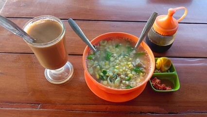 A bowl of steaming hot corn soup with meat and vegetables served with a glass of iced coffee, chili sauce and a side of lime.