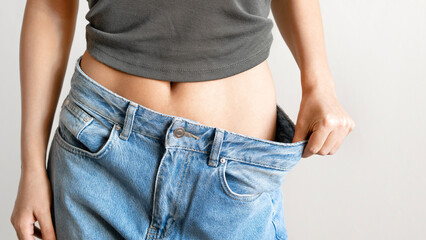 Diet and weight loss concept. Young woman in big jeans showing her slim body on light grey background. 