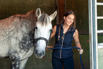 Smiling woman rider prepares to lead her horse out of the stable by pulling the harness rope