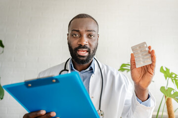 An African-American doctor holding a blister pack of pills and a clipboard explains medication instructions during online consultations. He appears professional and attentive.