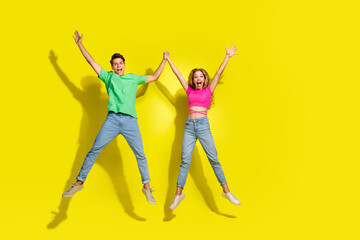 Full body portrait of two nice young partners hold raise arms jump empty space isolated on vibrant yellow color background
