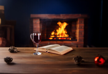 A glass of red wine sits on a wooden table next to an open book with a crackling fire in the background.