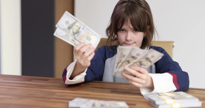 Smiling schoolgirl counts dollar banknotes sitting at desk. Cheerful child engages in game with fake money improving financial literacy slow motion