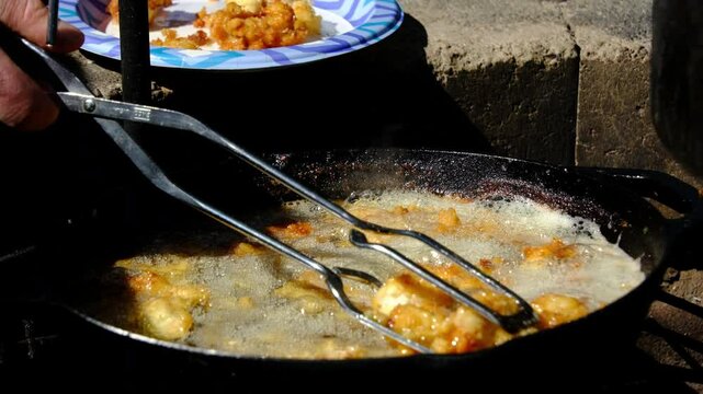 Slow motion and close up of browned fish nuggets being removed from pan after deep frying over fire pit at camp site