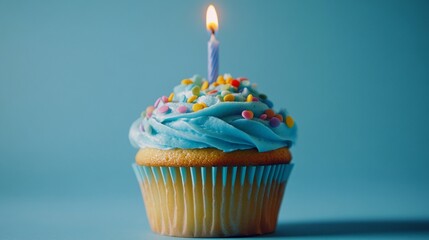 A single cupcake with blue frosting and colorful sprinkles, topped with a lit candle, against a light blue background.