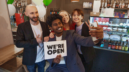 Diverse group of colleagues taking selfie together, holding we are open sign to celebrate launch of new business. Opening coffee shop, cafe. Concept of business, achievement, success, entrepreneurship