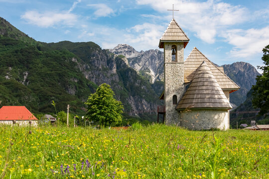 Picturesque stone church of St. George on blooming alpine meadow in calm village Theth, Northern Albania. Surrounded by pine tree forest and steep mountain slopes of Albanian Alps (Accursed Mountains)
