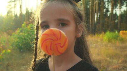 A girl in a carnival witch costume holds a sweet candy on autumn forest park on the sunset. Trick or treat for Halloween takes candy. Childhood, traditions and fun concept. Slow motion - Powered by Adobe