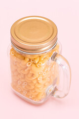Uncooked Chifferi Rigati Pasta in Glass Jar on Pink Background. Fat and Unhealthy Food. Scattered Classic Dry Macaroni. Italian Culture and Cuisine. Raw Pasta