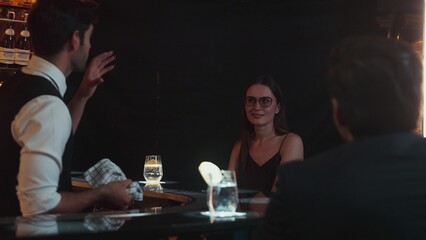 Focusing on beautiful woman guest sitting in special drink gin fizz at luxurious counter bar at vibrant nightlife party atmosphere. Smart bartender wiping glass while talking with customer. Vinosity.
