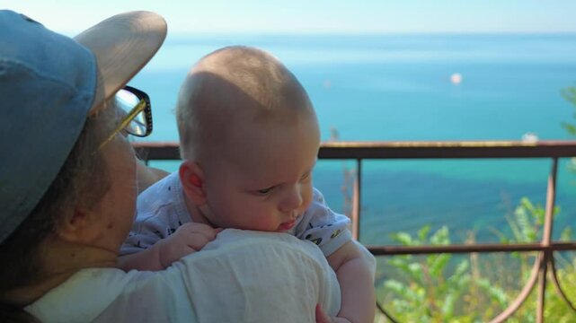 A woman in a baseball cap is holding a baby in a white jumpsuit, sitting on a bench by sea. Baby looks over her shoulder. Turquoise water stretches to horizon, metal railings frame serene seascape