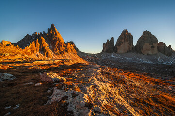 Obraz premium Majestic peaks of the Tre Cime di Lavaredo, Dolomites bathed in golden sunlight at dusk as the rugged landscape showcases nature’s beauty in Italy's renowned mountain range.