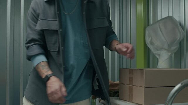 Caucasian man loading cardboard boxes, plastic container and suitcase on cart inside of modern metal-walled self-storage unit