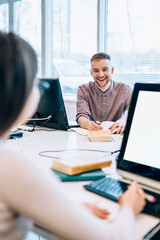 Cheerful students consulting while reading textbook and studying in library computer room