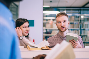 Positive multiethnic students talking while searching for textbooks in library