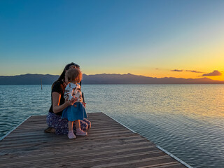 Caring mother holding toddler on wooden pier on calm Skadar lake at sunset in Shkoder, Albania. Scenic view of Dinaric Alps mountains, Southern Montenegro. Perfect family vacation. Child love emotion