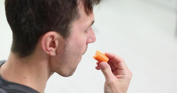 Man eats bright thin carrot chewing bit off piece with pleasure. Man enjoys taste of fresh vegetable carefully examining product in hand slow motion