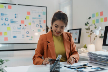 Young asian businesswoman working in a modern office analyzing charts and taking notes with documents and computers on her desk