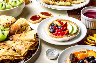Festive breakfast spread with crepes, fruit tart, and tea. Delicious breakfast with fresh fruit, pancakes, cookies, and tea. Colorful table setting with homemade pancakes, tart, and fruit.