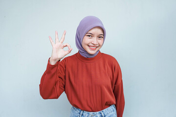 Beautiful smiling Asian woman in red sweater showing okay gesture demonstrates symbol of approval isolated over gray background.