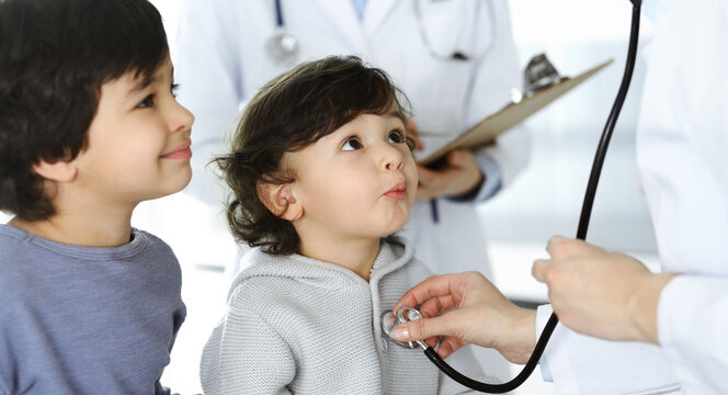 Woman-doctor examining a child patient by stethoscope. Cute arab toddler and his brother at physician appointment. Medicine concept
