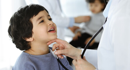 Doctor-woman examining a child patient by stethoscope. Cute arab boy and his brother at physician appointment. Medicine concept