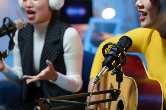 Two young women are recording music in a professional sound studio, one singing and the other playing acoustic guitar
