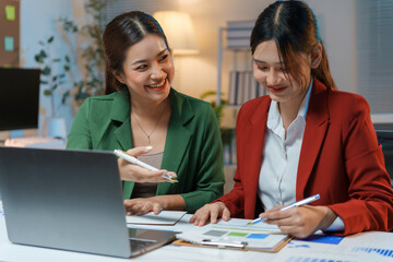 Obraz premium Two young businesswomen are working late in the office, collaborating on a project and analyzing financial data on a laptop