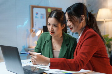 Fototapeta premium Two businesswomen are working late in the office, discussing a project and pointing at a laptop screen