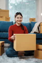 Young woman is sitting on the floor of her new apartment, surrounded by moving boxes, holding a cardboard box