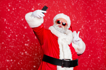 Portrait of his he nice attractive cheerful cheery positive flirty bearded Santa pouted lips taking making selfie having fun showing v-sign isolated over bright vivid shine red background