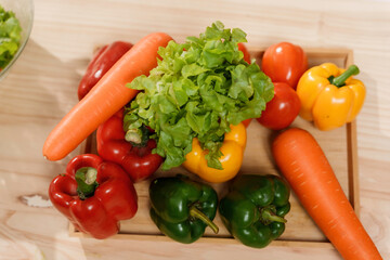 Fresh lettuce, carrots, bell peppers, and tomatoes lying on wooden cutting board in kitchen