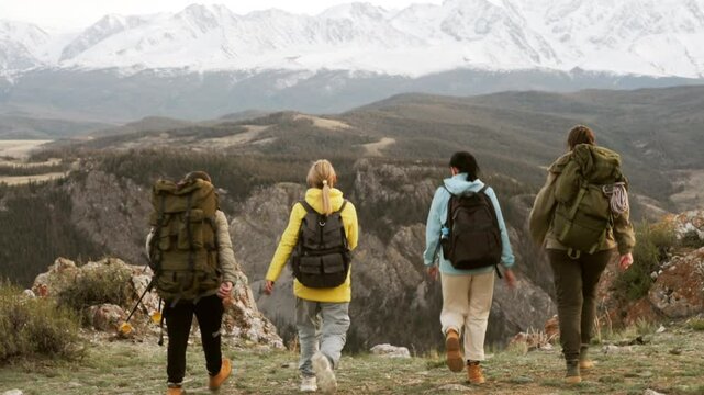 Back view at group of four hikers with backpack raising arms achieving success and top standing on mountain cliff. Motivation to travel together, active recreation on scenic peak