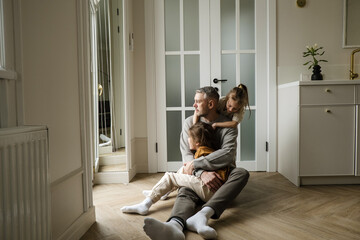 A father sits on the floor with his two daughters, surrounded by love and warmth. One daughter is nestled in his arms, while the other stands with her arm around his shoulder.