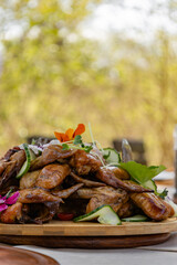 BBQ chicken wings garnished with microgreens, flowers and fresh cucumber slices. Beautiful serving on a wooden plate. Side view. Aesthetic outdoor dinner