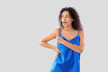 Young African-American woman in silk dress pointing at something on light background