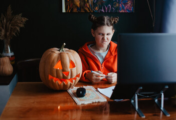 Caucasian happy girl orange color cloths near carved Halloween pumpkin studying online laptop