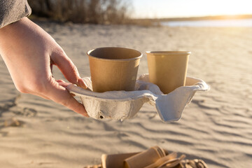Two eco-friendly paper cups on a paper tray, placed on a sandy beach by the river or sea at sunset. Perfect for branding mockups with space for text or design, ideal for promoting eco-conscious produc