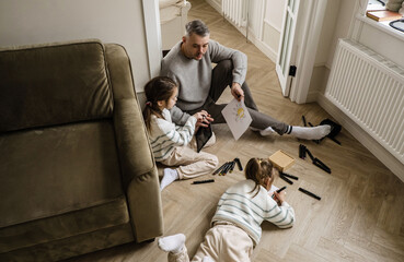 A father and his two daughters are enjoying a creative afternoon. The father is showing a drawing to his older daughter while she is using a tablet.