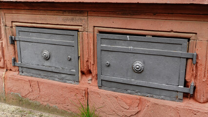 Two vintage access doors are set into a weathered brick wall, showcasing decorative hinges and a worn texture, highlighting the passage of time and architectural detail.