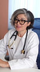 Caucasian doctor with gray hair, black glasses adjusts stethoscope on neck, wearing white coat, focused on patient discussion with attentive posture, exuding composed confidence. Attentive leadership.