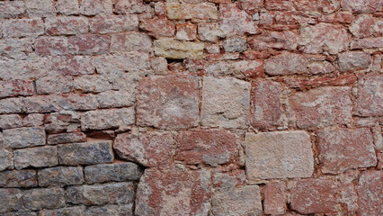 A weathered brick wall displays a mix of red, beige, and gray stones, showcasing a rich history through its uneven textures and color variations, located in an outdoor environment.