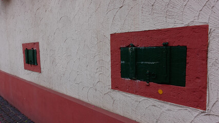 In a quaint village, green shutters are mounted on a textured white wall with red borders. The shutters add a touch of charm and historic character to the buildings exterior.