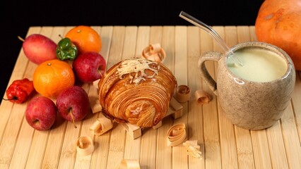 Croissant near ceramic mug filled with creamy vegetable beverage, fruits scattered on bamboo mat, vibrant autumn harvest scene highlighting fresh seasonal produce, rustic textures, natural light.