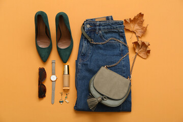 Stylish female bag with jeans, shoes, accessories and autumn leaves on orange background
