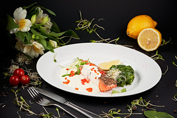 Plate with red fish, artistically presented on a black background framed by vegetables and flowers....