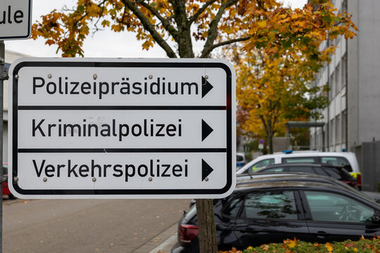 Ein Schild weist den Weg zu verschiedenen Abteilungen des Polizeipr&auml;sidiums, darunter die Kriminalpolizei und die Verkehrspolizei. Im Hintergrund sind geparkte Polizeifahrzeuge zu sehen.