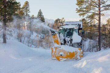 Snow plow tractor with rear-mounted snowblower cleaning a snowy mountain road, pushing snow high...