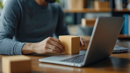 A man is working at a desk with a laptop and a cardboard box. He is holding the box in his hand and looking at the screen.