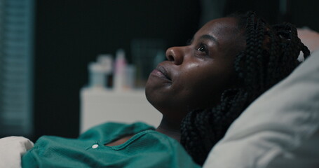 Young African patient lies in her hospital bed, lips pressed together in a contemplative...